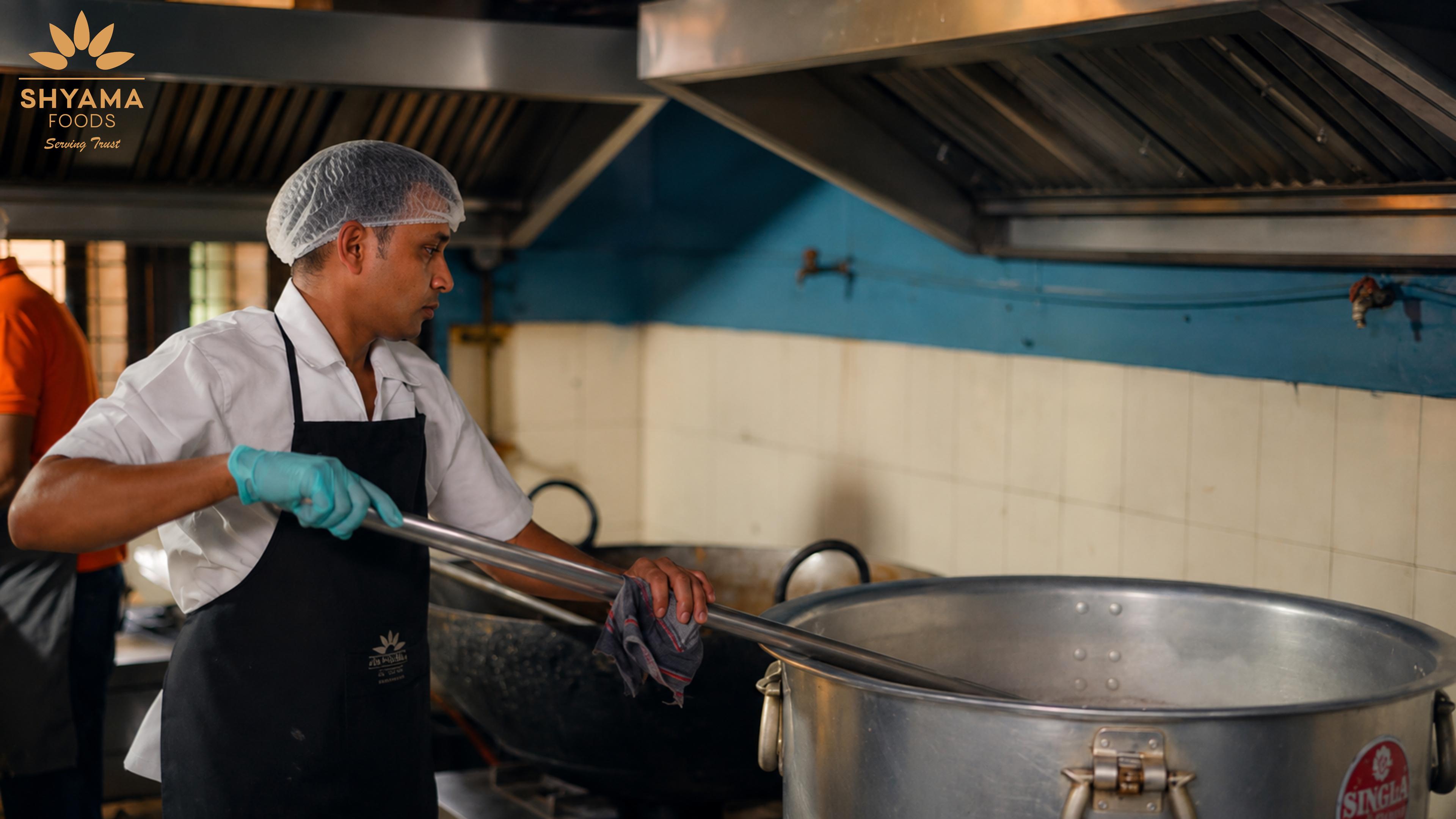 Chefs cooking a large batch of food in a big vessel at the Shyama Foods kitchen